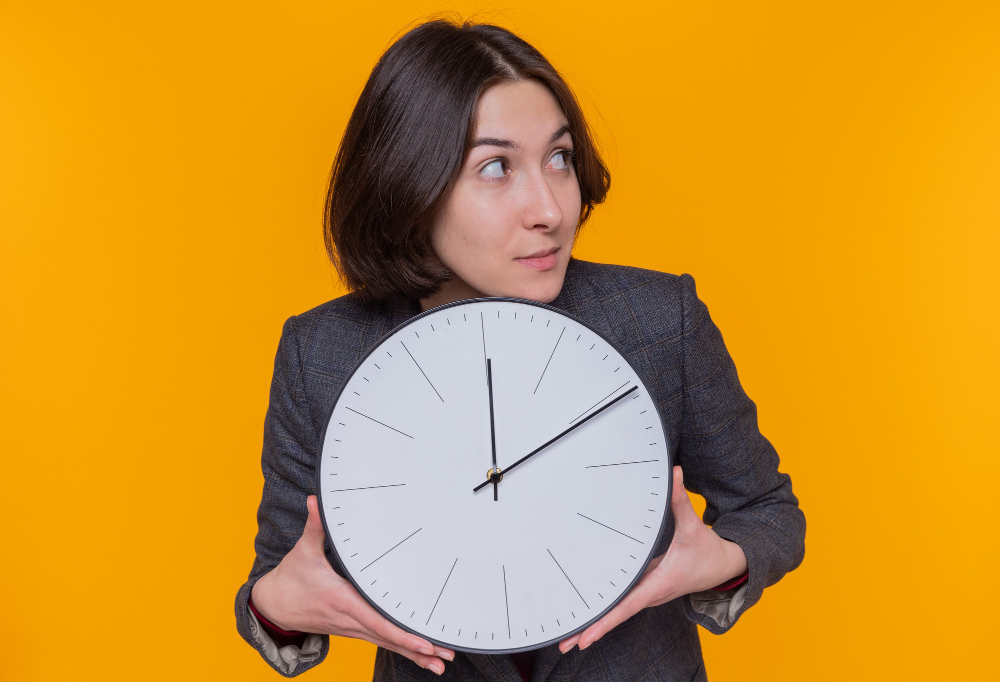 young-woman-with-short-hair-wearing-grey-jacket-holding-wall-clock-looking-aside-with-a-suspicious-expression-standing-over-orange-wall.jpg